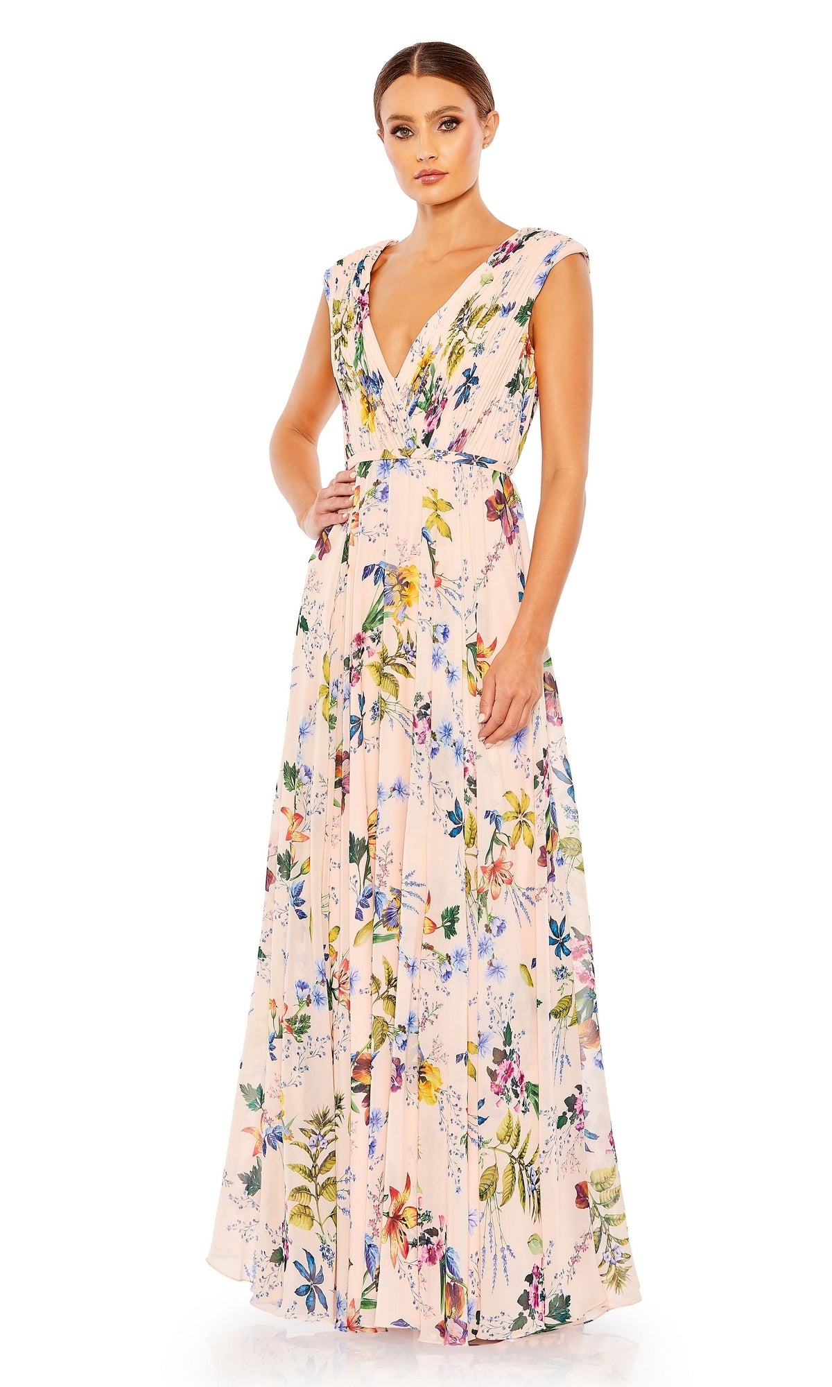 A woman wears the Mac Duggal 55829 Long Floral-Print Formal Dress, a sleeveless light pink V-neck gown with vibrant floral designs, shown standing against a white background.